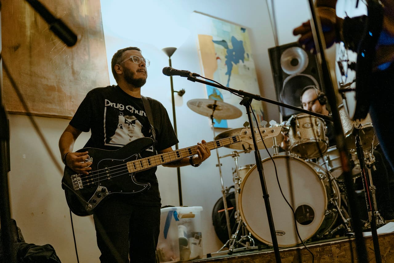 A musician playing bass in a rehearsal room, with a drum kit behind